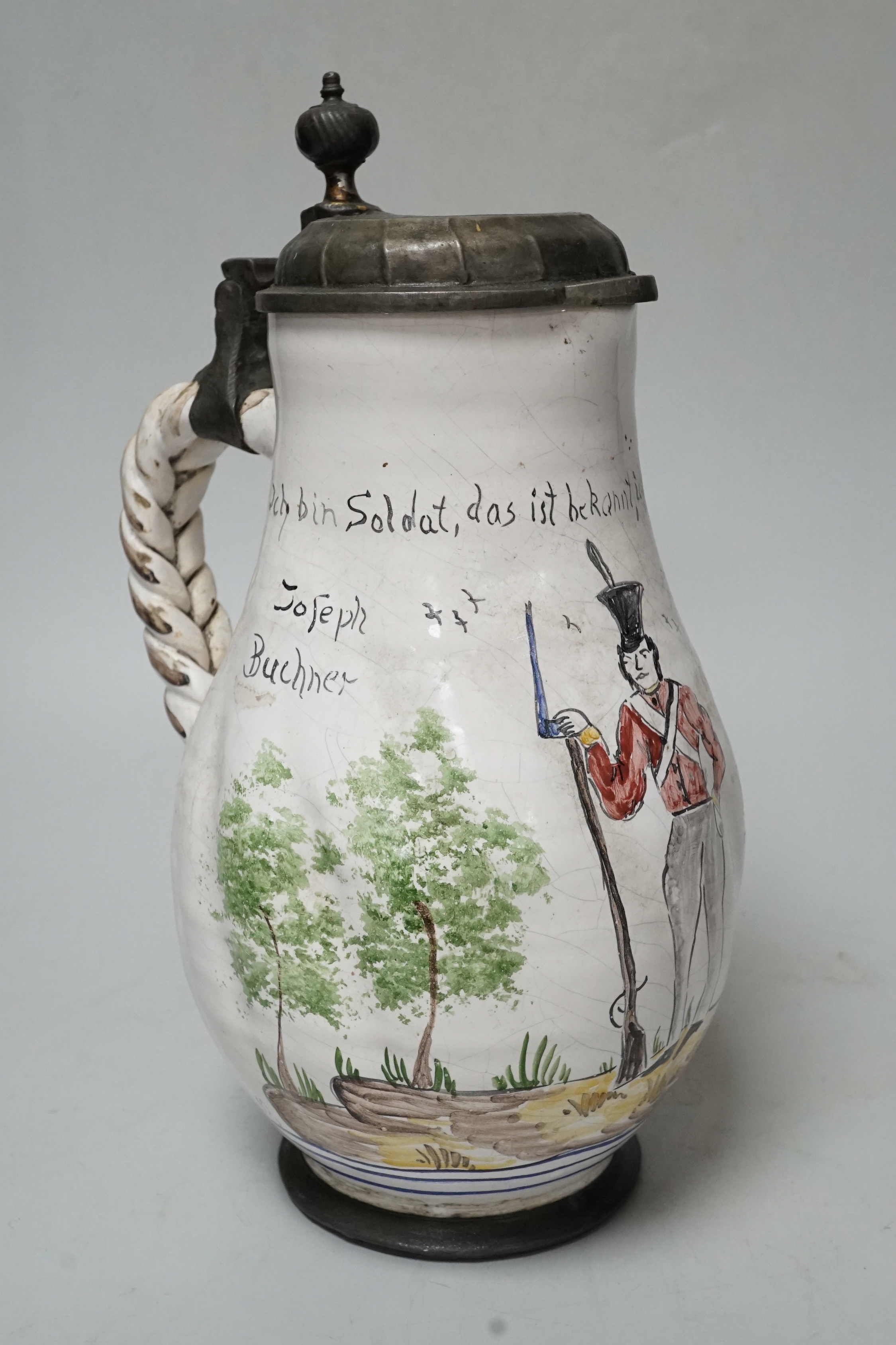 A large 18th century German faience pewter lidded jug, baluster shaped, with a naive painting of a soldier, inscribed ‘’Soldat, das ist bekqnnt, ich streite fur das vaterland’’ (Soldier, that is known for that fatherland